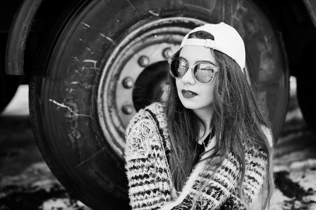 Brunette stylish casual girl in cap and sunglasses sitting against truck wheels.